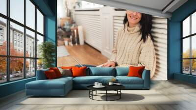 Indoor shot of smiling young woman looking to the camera and shopping online on laptop in cozy Christmas interior. Pretty female sitting on the floor, bying on winter sales via computer using wireless Wall mural