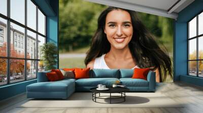 Outdoor close-up portrait of a beautiful young brunette woman smiling broadly and looking to the camera, posing against nature background in the park. Pretty female walking in the park. Wall mural