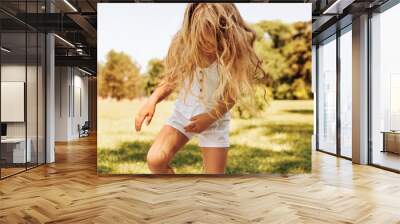 Outdoor image of smiling adorable little girl with long blond hair playing at nature background. Happy child enjoying summer time in the park. Cheerful kid have activity in in the forest on sunlight Wall mural