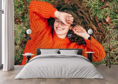 Overhead image of a joyful young woman wearing a knitted orange sweater lying on the grass. The pretty female has a positive expression, resting in the park. Wall mural