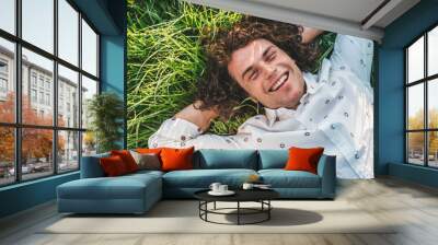 People, lifestyle. Outdoor close up portrait of happy handsome young male smiling with white toothy smile lying on the green grass in the park, looking at the camera with copy space for advertising. Wall mural