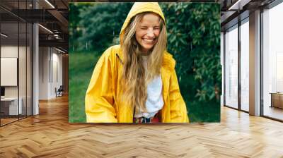 Playful young blonde woman smiling wearing yellow raincoat during the rain in the park. Cheerful female enjoying the rain outdoors. A happy woman enjoying the rainy weather. Wall mural