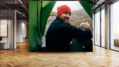 Portrait of a view from tent entrance of young traveler man smiling and relaxing in mountains near to bonfire. Rear view of hiker male sitting in the camping tent, drinking hot beverage. Travel people Wall mural