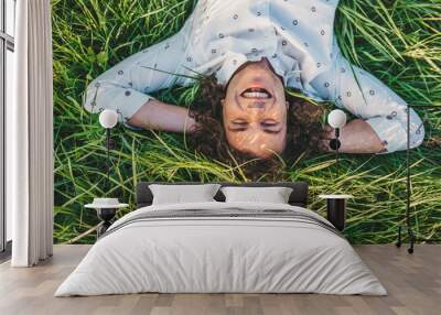 Portrait of happy handsome young male with curly hair smiling and lying with hands behind head on the green grass in the park looking at camera.Copy space for advertising. People and lifestyle concept Wall mural