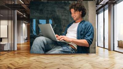 Portrait of happy young man with curly hair using laptop for working online. Smart freelance man chatting online with clients. Handsome male reading news, typing on his laptop computer in the office Wall mural