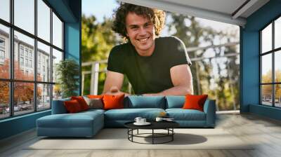 Portrait of smiling cheerful young man wears black t-shirt and wristwatch on the city street. Happy male with curly hair posing for advertisement with copy space outdoor. People concept Wall mural