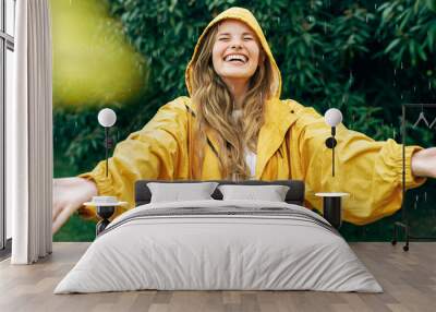 Positive young blonde woman smiling wearing yellow raincoat during the rain in the park. Cheerful female enjoying the rain outdoors. A beautiful woman catching the raindrops with arms wide open. Wall mural