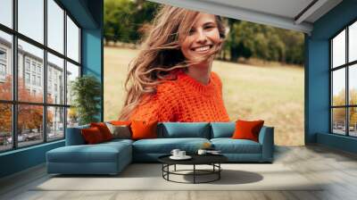 Positive young woman wearing a knitted orange sweater posing on nature background. A cheerful female has a joyful expression during walking in the park. Wall mural