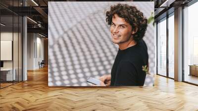 Rear view image of cheerful man in black t-shirt reading text message on his mobile phone on the street. Young Caucasian male with curly hair looking to the camera with copy space for text. Wall mural