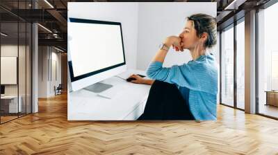 Side view image of freelancer businesswoman using a desktop computer in an office. Young blonde woman sits indoors at home working on the computer with a blank screen for your text message or content. Wall mural