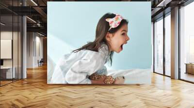 Side view of happy little girl in pajama has an amazed expression afterwake up in the bed, isolated on light blue background. Kid has joyful expression in the morning. Wall mural