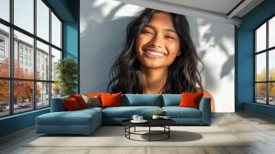 Smiling woman with long dark hair and glowing skin standing in soft light with a white wall and tree shadows behind her. Wall mural