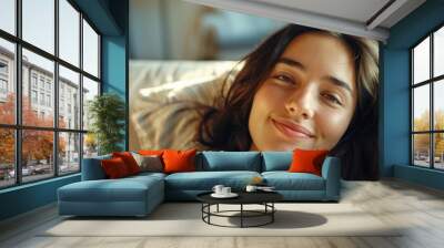 Smiling young woman in casual attire resting on a stylish sofa, surrounded by a bright and modern home setting. Wall mural