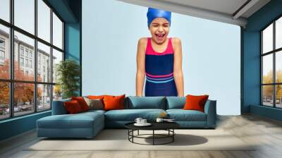 Studio image of a little funny happy girl in a swimsuit, isolated over blue studio background. A joyful wet kid in a swimsuit and swim cap smiling broadly during swimming training. Wall mural