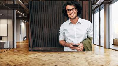 Successful businessman in eyewear smiling broadly posing outdoors. Happy male entrepreneur messaging on mobile phone. Smart guy in casual wears spectacles with curly hair walking outside after work Wall mural