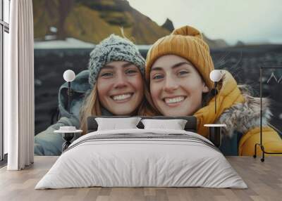 Two smiling women in winter clothing with beanies and jackets posing for a selfie in a snowy landscape with a mountain in the background. Wall mural