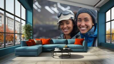 Two smiling women one wearing a blue jacket and a beanie the other in a grey jacket and a cap standing outdoors with a mountainous background possibly during a hike or adventure. Wall mural