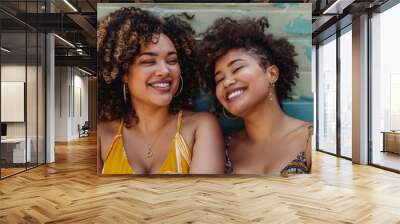 Two women with curly hair smiling and leaning against a blue wall with peeling paint wearing summer dresses and hoop earrings exuding joy and friendship. Wall mural