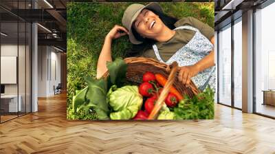 View from above of a gorgeous female gardener lying on the grass with a basket with freshly picked ecological vegetables at the farm. A farmer woman resting after picking the harvest in the garden. Wall mural