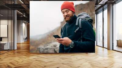 Young bearded man smiling and sending messages for his family from his cellphone, during hiking in mountains. Traveler bearded man in red hat using mobile phone application. Travel and lifestyle Wall mural