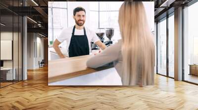 A young barista in a black apron smiles as he brings a cup of co Wall mural