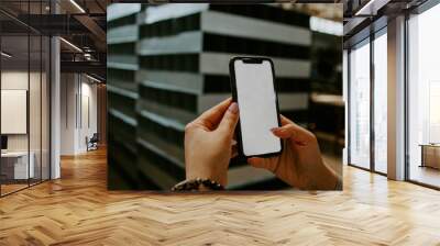 A hand holds a white mobile phone with an empty white screen on the background of the factory Wall mural