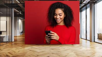 Beautiful young smiling woman of african beauty looks at phone wearing red sweatshirt isolated on red background with space for text or inscriptions.generative ai
 Wall mural