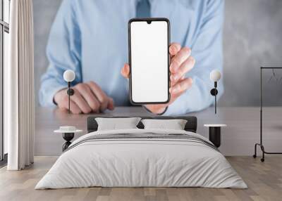 Businessman in a blue shirt at workplace at the table holding a mobile phone, smartphone with a white screen. Screen facing the camera. Mock up.Concept of technology, connection, communication. Wall mural