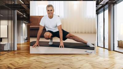 Elderly man doing side lunge exercise. Senior male athlete with fit body warming up, doing leg stretch workout at home. Take care of yourself Modern light room on background. Elderly sport concept. Wall mural