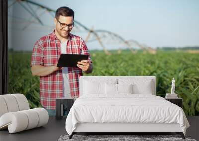 Happy young farmer or agronomist in red checkered shirt using tablet in corn field. Irrigation system in the background. Organic farming and food production Wall mural