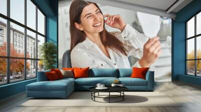 Happy young woman smiling checking out her perfect healthy teeth in the mirror, sitting in a dental chair at the dentist office Wall mural