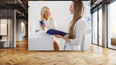 Medical worker consults patient in white hall of the hospital. Patient senior blond woman standing near the reception and holding a brochure with beauty treatments Wall mural