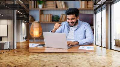 Overjoyed happy indian guy, lucky winner, sitting at desk watching virtual lottery results on laptop shaking hands. Eastern student got perfect exam grades, job proposal offer. Wall mural