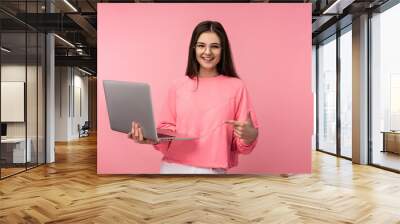 Photo of attractive lady in glasses, smiles and has online meeting, holds laptop and works online. wears casual pink t-shirt white pants isolated pink color background. Online study. Wall mural