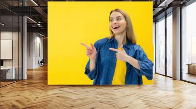 Positive woman smiles positively recommends using blank space indicates at upper left corner dressed in casual wear over vivid yellow background shows direction or way Wall mural