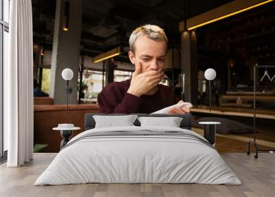 Spoiled food in the restaurant. Dissatisfied young man in a cafe covers his mouth with a hand disgusted with a taste of food. A meal on the table in front of him. Bad customer service concept. Wall mural