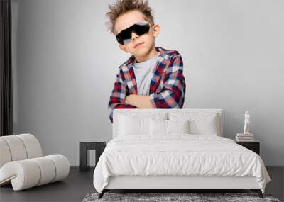 A handsome boy in a plaid shirt, gray shirt and jeans stands on a gray background. The boy in the black sunglasses. The boy folded his arms over his chest Wall mural