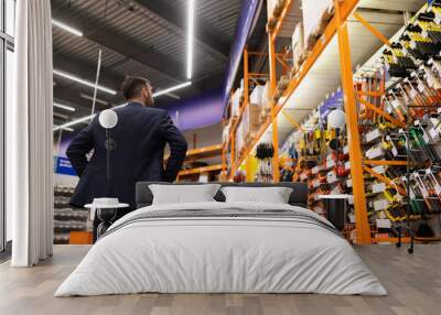 a man in a hardware store in front of shelves with saws and hammers stands with his hands folded on his sides with his legs wide apart, photo from the back, the concept of choosing goods Wall mural