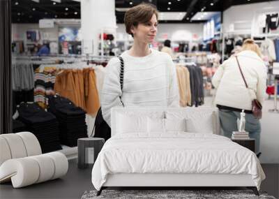 A young beautiful and attractive brunette woman with a short hair in a casual and stylish white sweater chooses fashionable jeans and denim clothes in a store in a shopping mall. Shop concept Wall mural