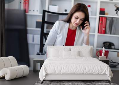 A young girl sitting in the office at the computer Desk and talking on the phone. Wall mural