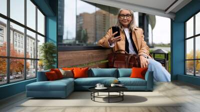 an older woman with a mobile phone sits on a bench outside Wall mural