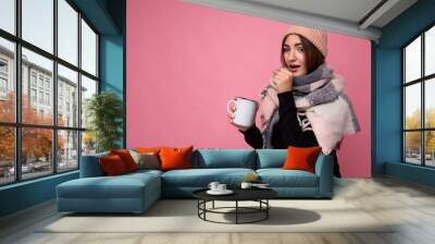 Beautiful sick young brunet woman wearing black sweater hat and warm scarf isolated over pink background holding paper mug for mockup drinking hot tea and looking at camera and caughing Wall mural