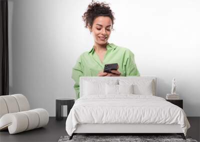 charming young brunette curly woman with a ponytail hairstyle is dressed in a green shirt and jeans typing a message on the phone Wall mural