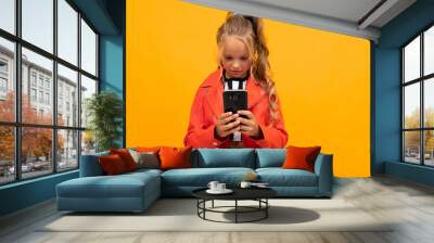 Cheerful girl in t-shirt and trousers smiles with her smartphone isolated on yellow and orange background Wall mural