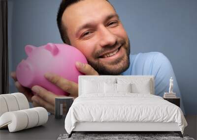 Closeup of delightful positive smiling young handsome brunet unshaven man with beard with sincere emotions wearing casual blue t-shirt isolated over blue background and holding pink piggy box. Money Wall mural