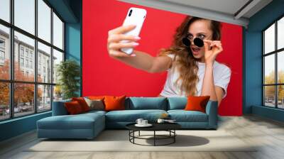 Closeup of happy amazing beautiful young woman holding mobile phone taking selfie photo using smartphone camera wearing everyday stylish outfit isolated over colorful wall background looking at device Wall mural