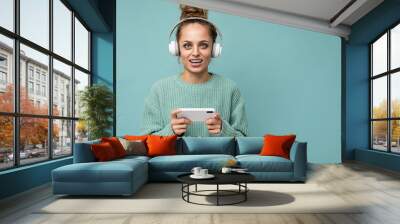 Closeup photo of beautiful positive happy young woman wearing blue sweater isolated over blue background wearing white bluetooth wireless earphones and listening to music and using mobile phone Wall mural