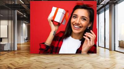 Closeup portrait photo of beautiful happy smiling young brunet woman isolated on red background wall wearing white casual t-shirt and stylish red and black shirt holding white gift box with red ribbon Wall mural