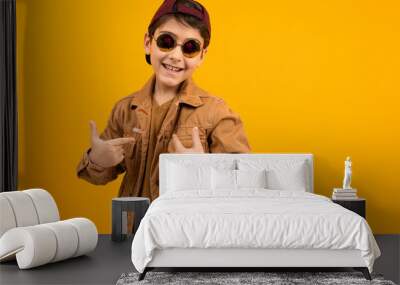 European teenager boy in a casual stylish brown denim jacket and stylish glasses shows himself with his hand on a yellow background with copy space Wall mural