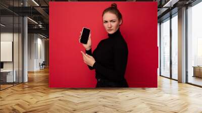 Photo of attractive serious young brunette woman wearing black sweater standing isolated over red background showing mobile phone with empty screen for mockup looking at camera Wall mural